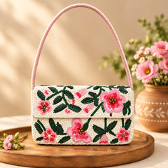 A shoulder bag with a flap closure, featuring a pink floral and green leaf beaded pattern on a light beige base